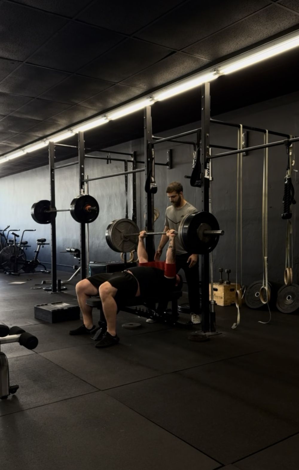 A coach standing behind a member on the bench press, hands ready to spot, as the member presses a loaded barbell
