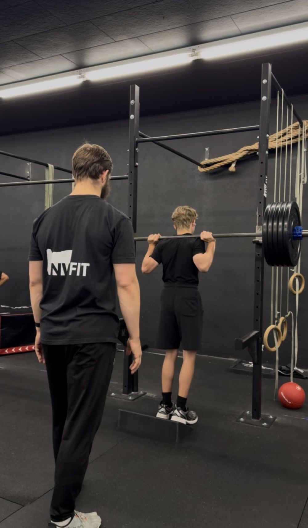 A coach in an NW FIT shirt watching an athlete's form as he holds a barbell at his shoulders while standing on a slant board