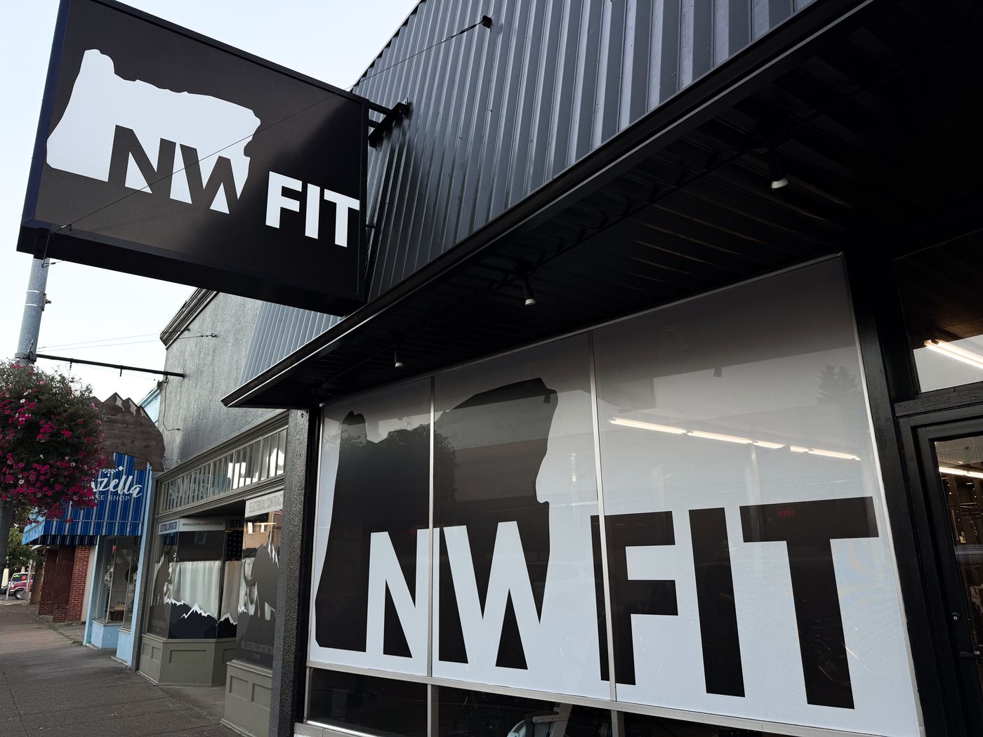 The NW FIT storefront on Main Street: a black hanging sign and large front windows printed with the NW FIT logo, which is shaped like the state of Oregon